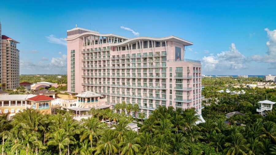 SLS Baha Mar in Nassau, Bahamas tall, pink modern hotel building with a wavy rooftop stands surrounded by lush palm trees. The sky is clear, creating a serene atmosphere.