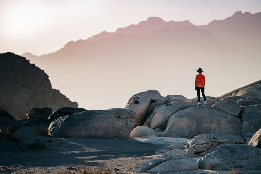 Person in orange shirt on rocky landscape during dawn or dusk, with mountains and pastel sky.