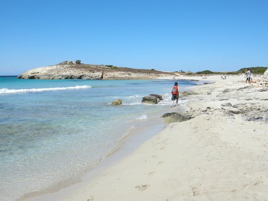 Tourists strolling on Son Bou Beach, Menorca, with soft sand and ocean waves in the background.