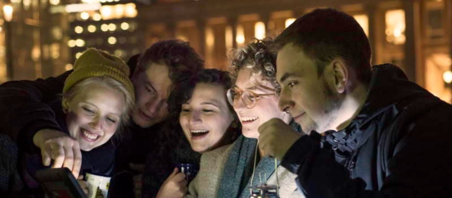 Group of young people gathered around a cell phone, engaged and discussing something exciting at TeamEscape SPACE ESCAPE Ulm.