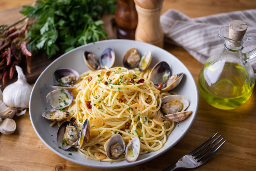 Bowl of spaghetti alle vongole with clams, garlic, and herbs sits on a wooden table. Olive oil, parsley, and garlic add a fresh, rustic vibe.