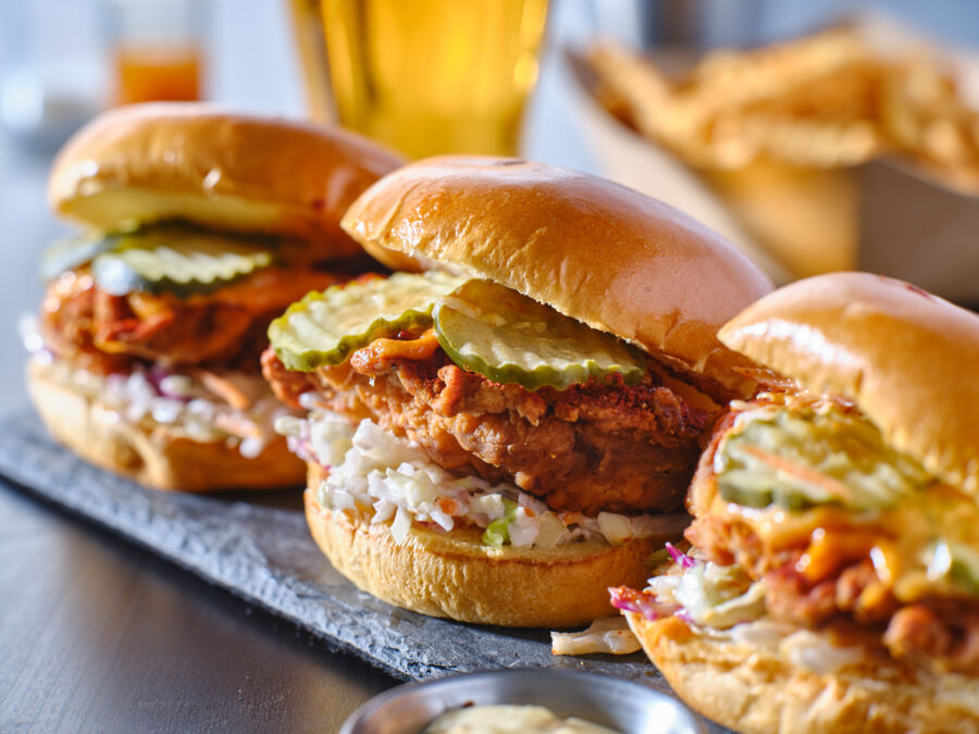 Three crispy chicken sliders on brioche buns, topped with pickles and coleslaw, sit on a gray slate with a side of sauce, exuding a delicious, appetizing vibe.