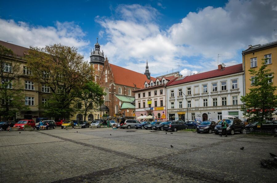 European town square with cobblestones, parked cars, historic architecture, church, and greenery.