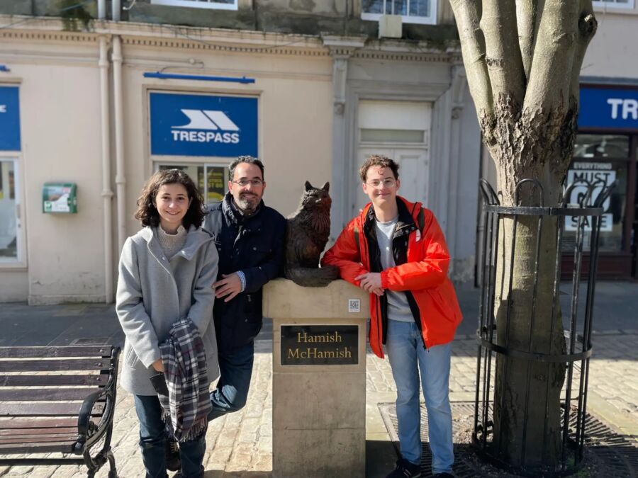 Three people smile for a photo in front of a statue during the St. Andrews Town Guided Private Walking Tour.
