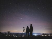 Photographers observing starry sky with tripod in serene night, city glow in background.