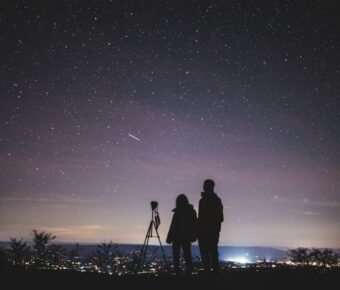 Photographers observing starry sky with tripod in serene night, city glow in background.