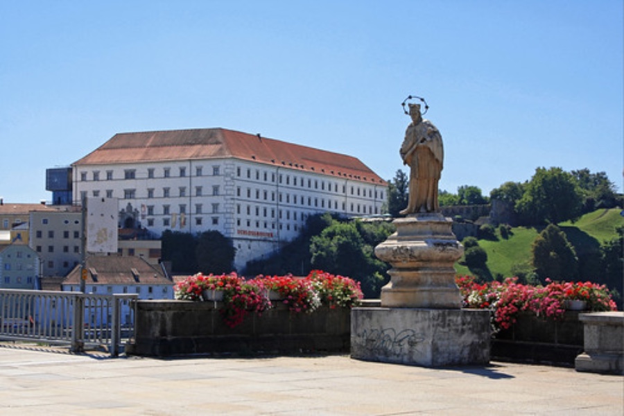 Stone statue on terrace overlook with red‑tiled historic building and flowering balustrade.