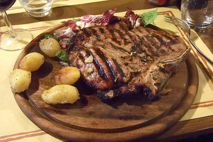 Grilled T-bone steak with roasted potatoes, fresh greens, and red wine on rustic platter.
