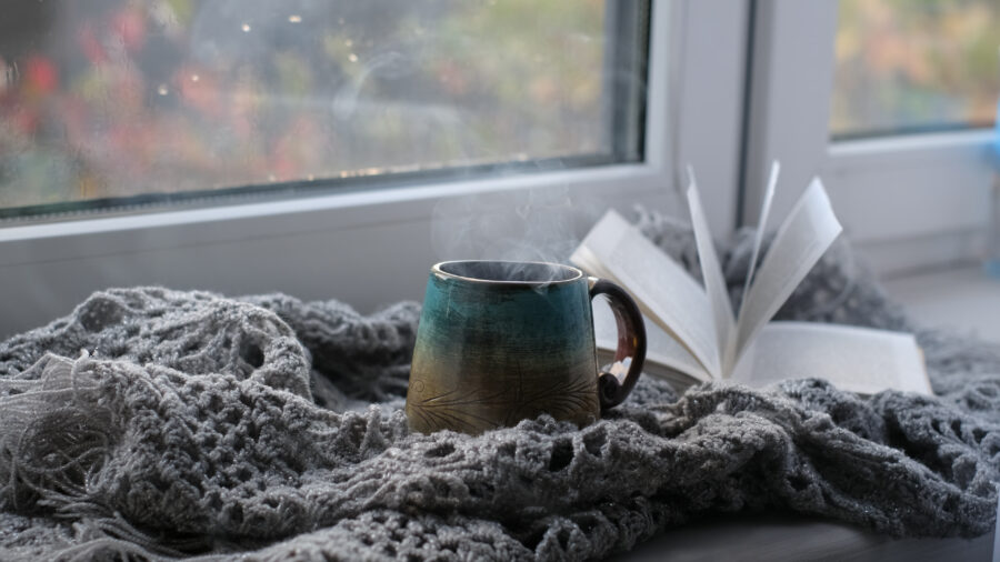 Steaming mug sits on a soft, gray knitted blanket beside an open book on a windowsill, creating a cozy and serene atmosphere.