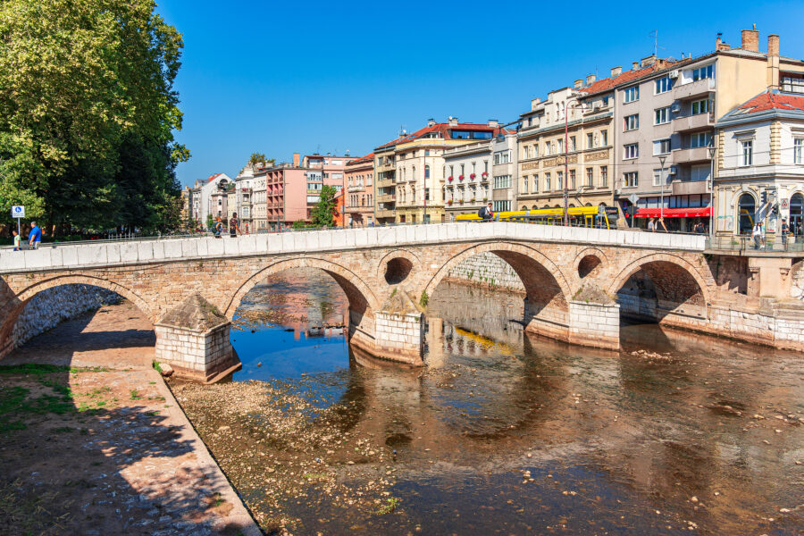 Historic stone arch bridge over shallow river in European city center with riverside buildings.