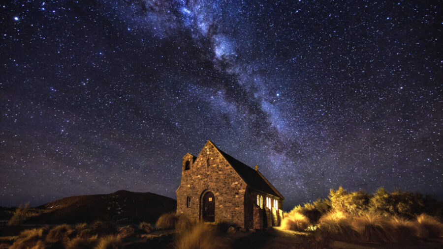 Stone chapel under Milky Way night sky with glowing doorway, stars, and rugged landscape.