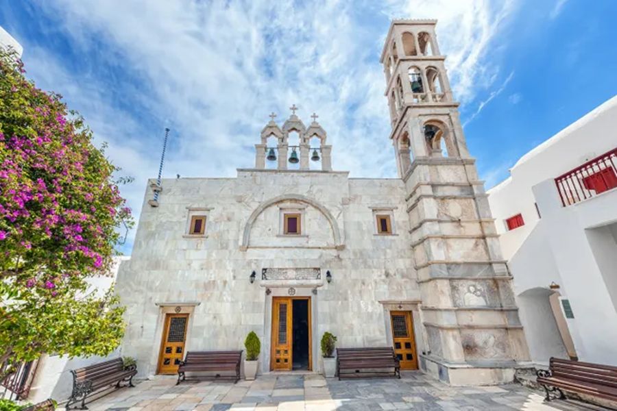 Church with light stone facade, ornate entry, bell tower, benches, greenery, and blooming bougainvillea.