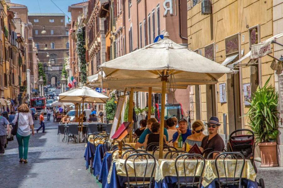 Italian cobblestone street cafe with outdoor umbrellas and patrons near Borgo Pio, Rome.