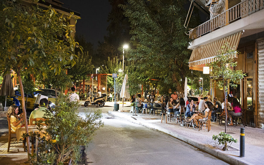 Nighttime street café with outdoor dining, people socializing under trees and warm lights.
