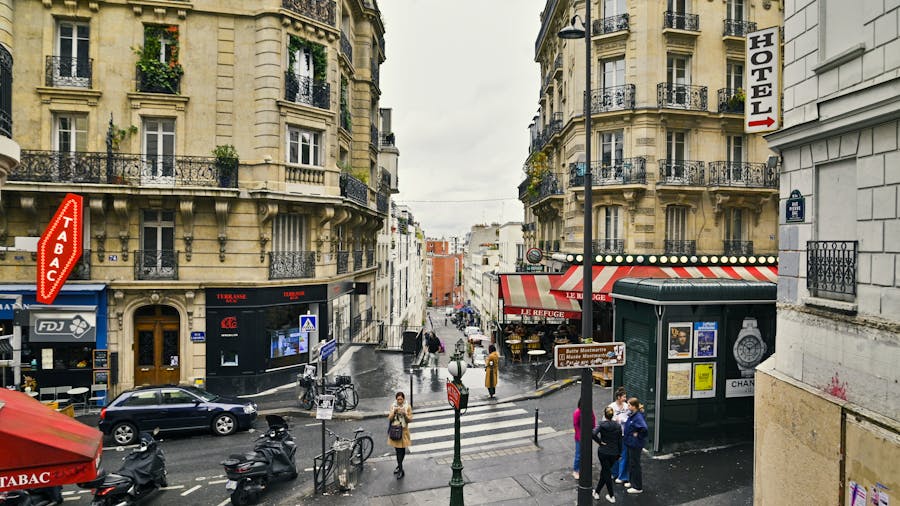 Rainy European street corner with cafe, wrought-iron balconies, zebra crossing, parked scooters.
