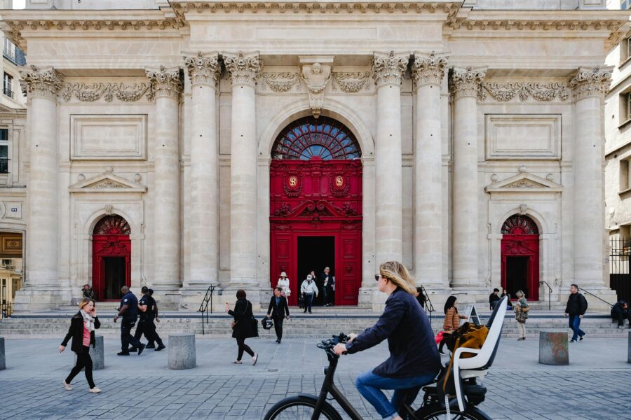 Neoclassical building with red doors, urban pedestrians, bikers, lively street scene.