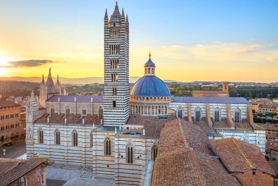 Duomo di Siena striped cathedral with dome and bell tower over terracotta roofs at sunset.