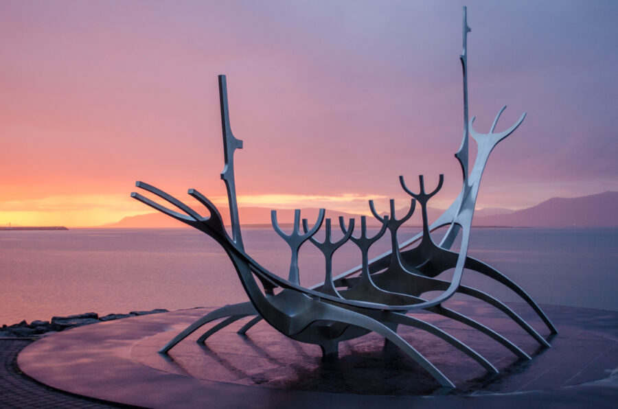 Modern boat sculpture with abstract, skeletal design set against a pink and orange sunset by a calm sea, conveying serenity and innovation.