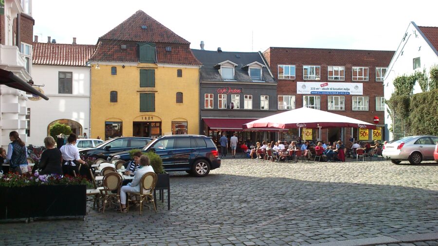 Sunlit cobblestone square with outdoor café seating, terraces, and people in a European Latin Quarter.
