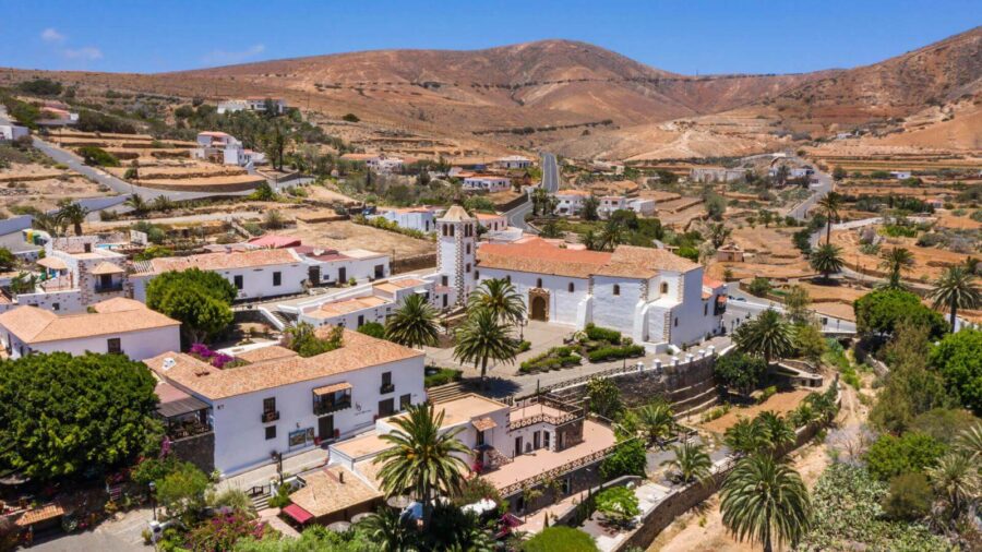 Sunlit Betancuria village with white church, palm trees, terraced hills, and arid Canary Islands landscape.