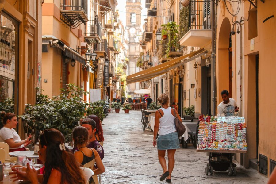 Sunlit Mediterranean cobblestone street with outdoor cafes and distant bell tower.