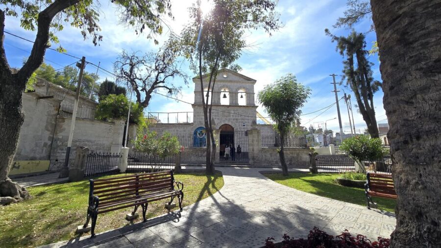 Sunlit town plaza with benches, trees, and historic stone San Lázaro church in Mexico.
