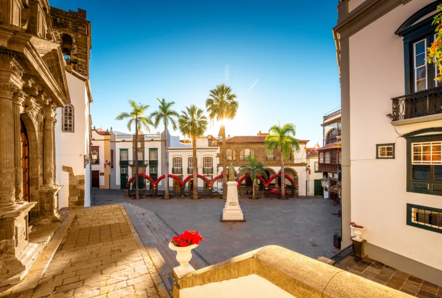 Santa Cruz de la Palma sunlit plaza with monument, lined palm trees, historic white buildings.