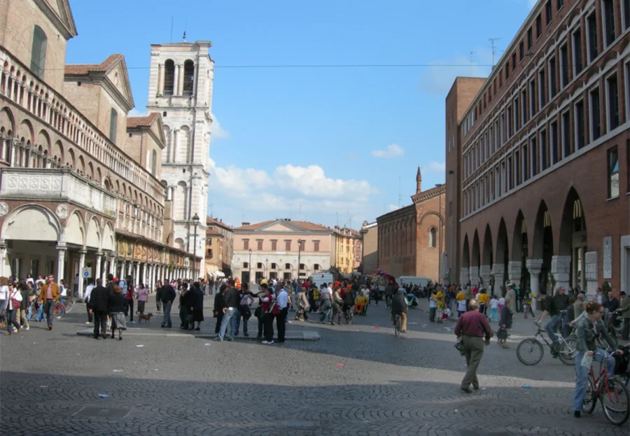 Sunlit Ferrara, Italy town square with cobblestone plaza, bell tower, arcades, and people.