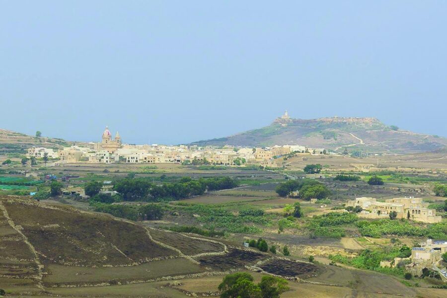 Gozo rural village with stone houses, terraced fields, and church dome in sunlit valley.