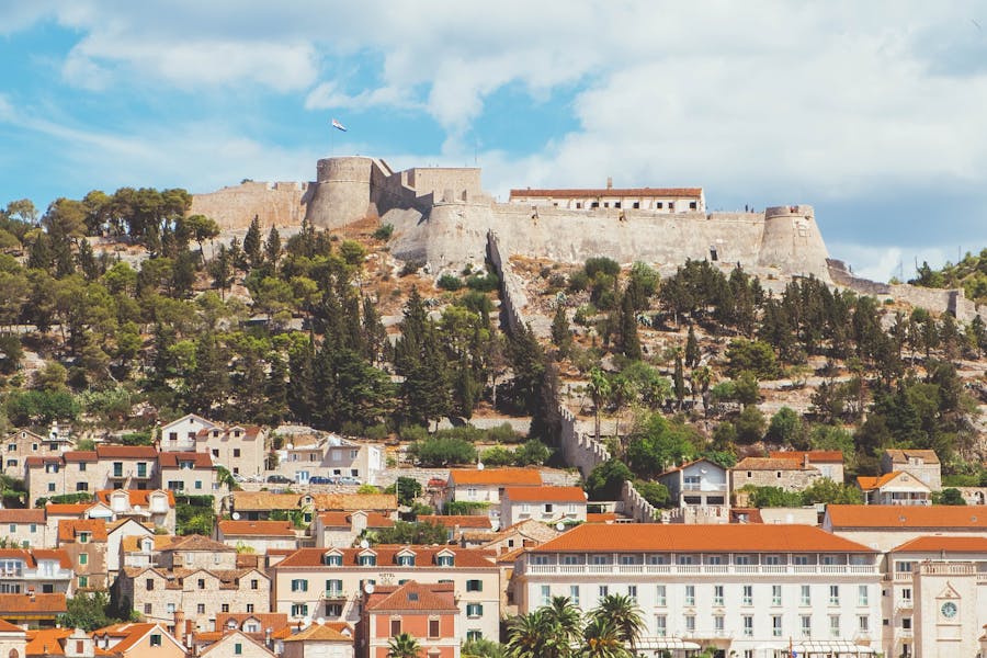 Sunlit Hvar coastal town with terracotta roofs and historic stone fortress on hillside.