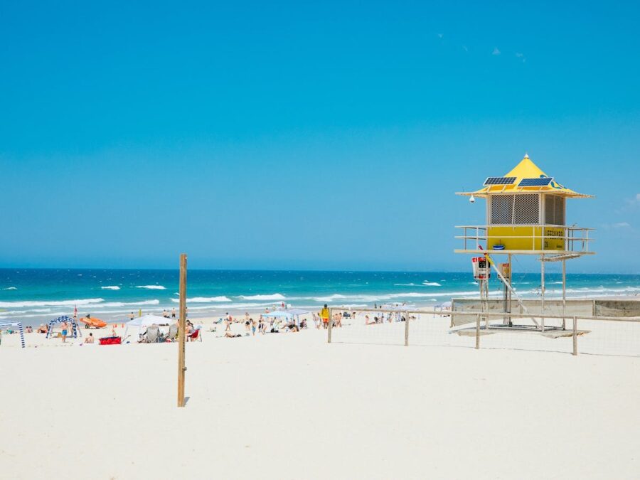 Sunny Kurrawa Beach with yellow lifeguard tower, blue ocean waves, umbrellas, and summer tourists.