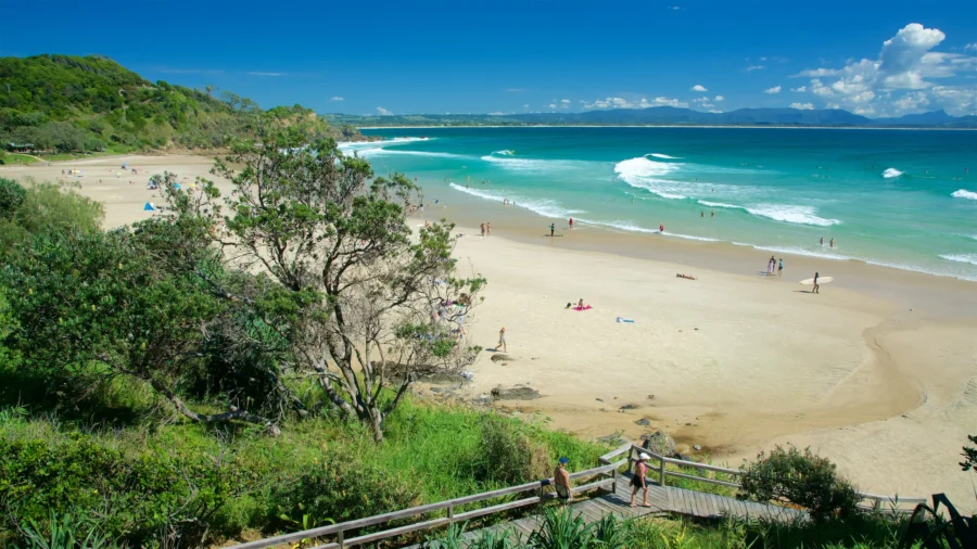Sunny Wategos Beach coastal boardwalk with turquoise ocean waves, white sand, headland, and beachgoers.