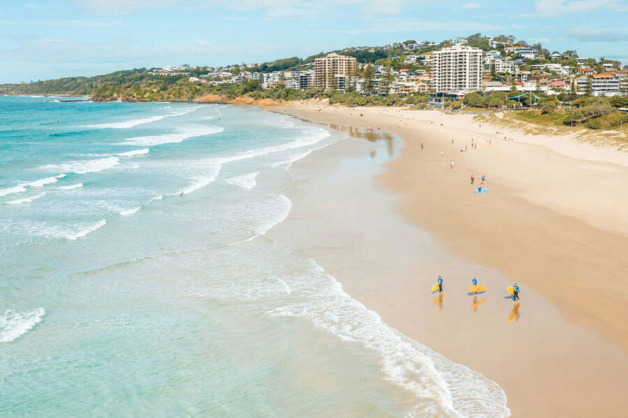 Sunny Coolum Beach coastal resort with surfers, gentle waves, golden sand, and oceanfront apartments.