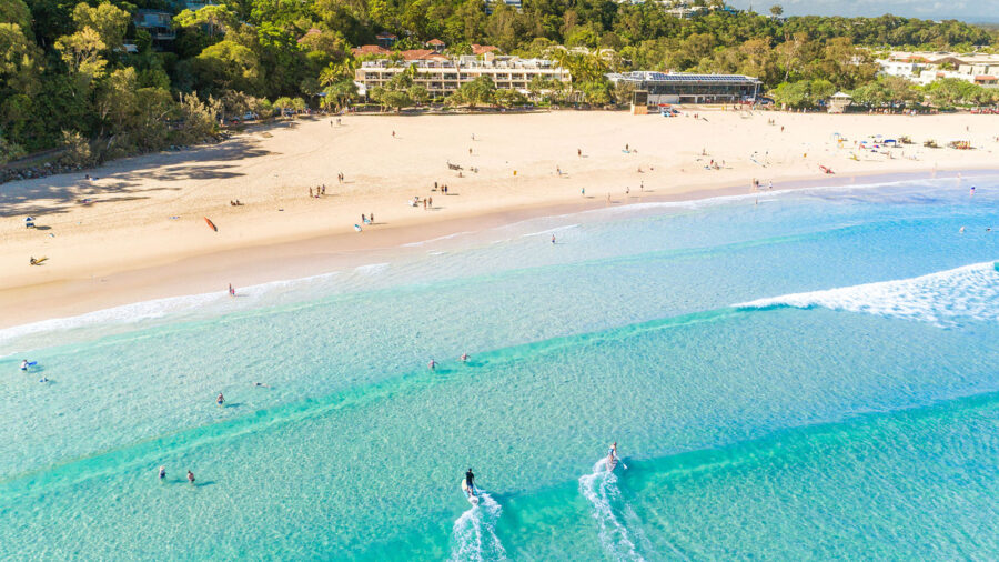 Aerial view of Noosa beach resort with turquoise ocean, surfers, golden sand, and coastal hotels.