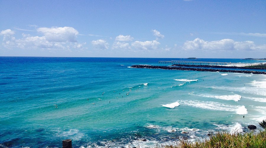 Surfers riding turquoise waves near pier and breakwater on sunny Coolangatta Beach coastline.