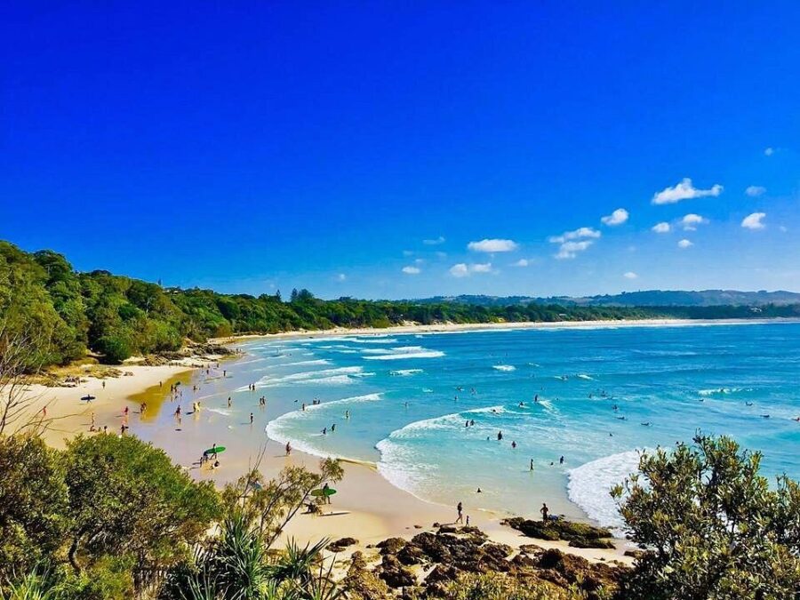 Scenic Belongil Beach coastline with turquoise ocean waves, golden sand, swimmers, surfers, rocks, and forest.