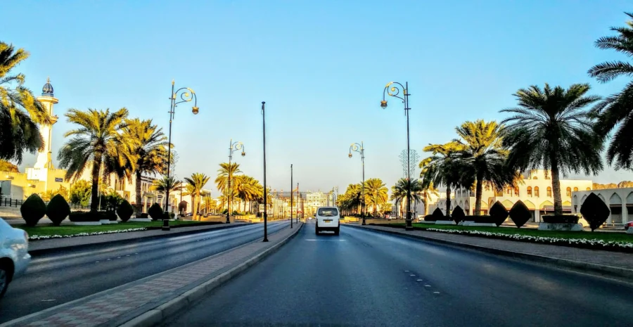 Sunny Al Ghubrah boulevard with palm trees, mosque minaret, white van and landscaped median.