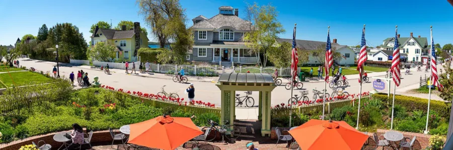 Sunny Mackinac Island street with cafes, American flags, cyclists, historic inns, and blooming tulips.