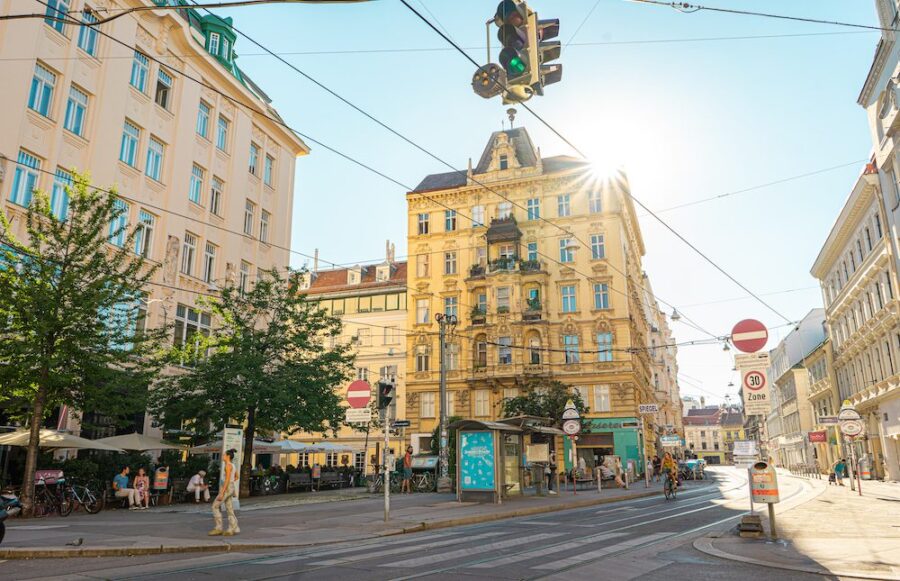 Sunny urban intersection with historic yellow building, tram tracks, cafes, and cyclists.