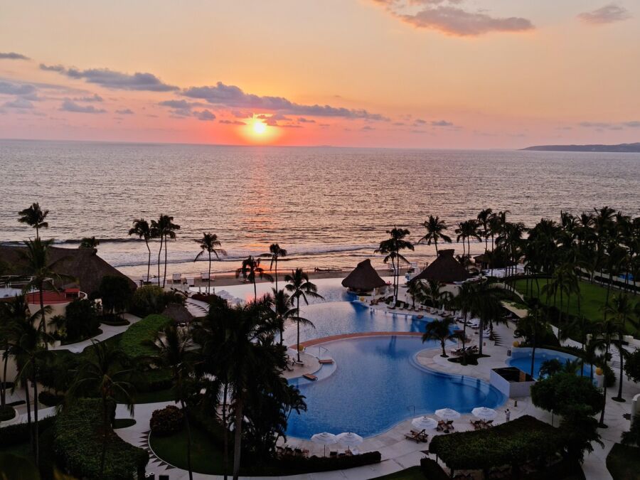 Tropical resort sunset with ocean, palm trees, pool, and colorful sky.