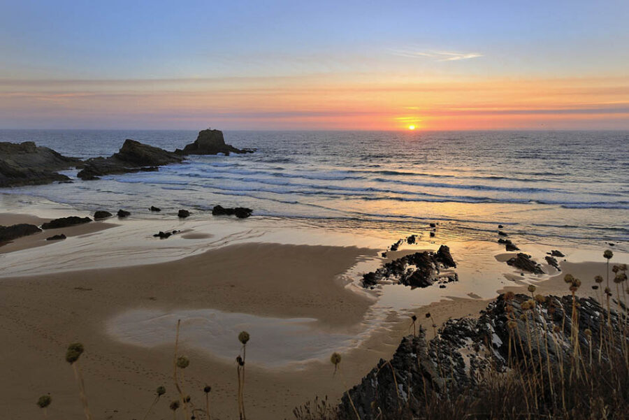 Zambujeira do Mar sunset beach with rocky coastline, waves, and golden ocean reflection.
