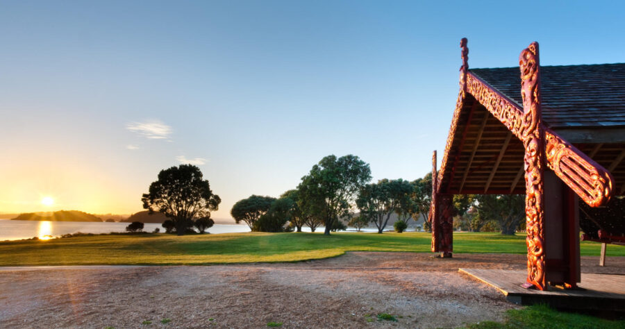 Sunset over Waitangi Treaty Grounds with Māori meeting house, carved wharenui, trees and coastal landscape.