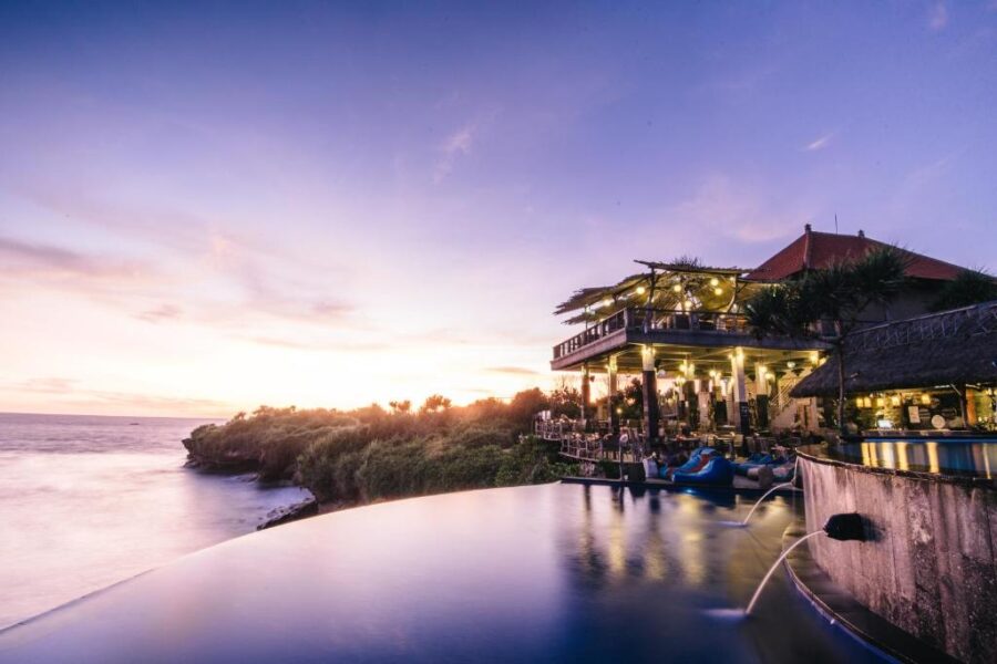 Luxury oceanfront infinity pool resort at sunset with cliffside restaurant, tropical foliage and glowing lights.