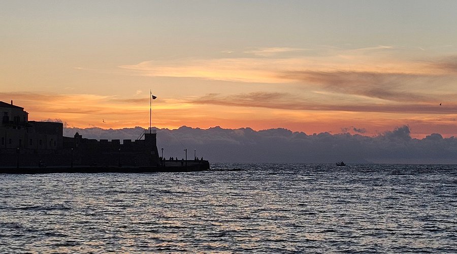 Serene coastal sunset with vibrant sky, calm water, silhouetted structure, and small boat.