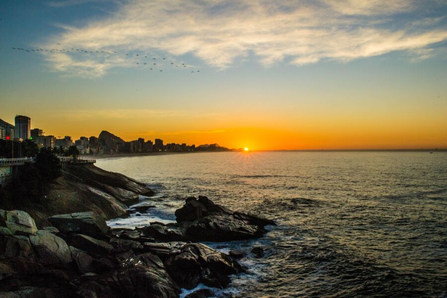Sunset over ocean with city skyline, rocks, colorful sky, and bird silhouettes.