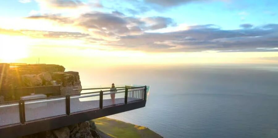 Glass-floored cliff deck with stunning ocean view, sunset sky, and rugged terrain.