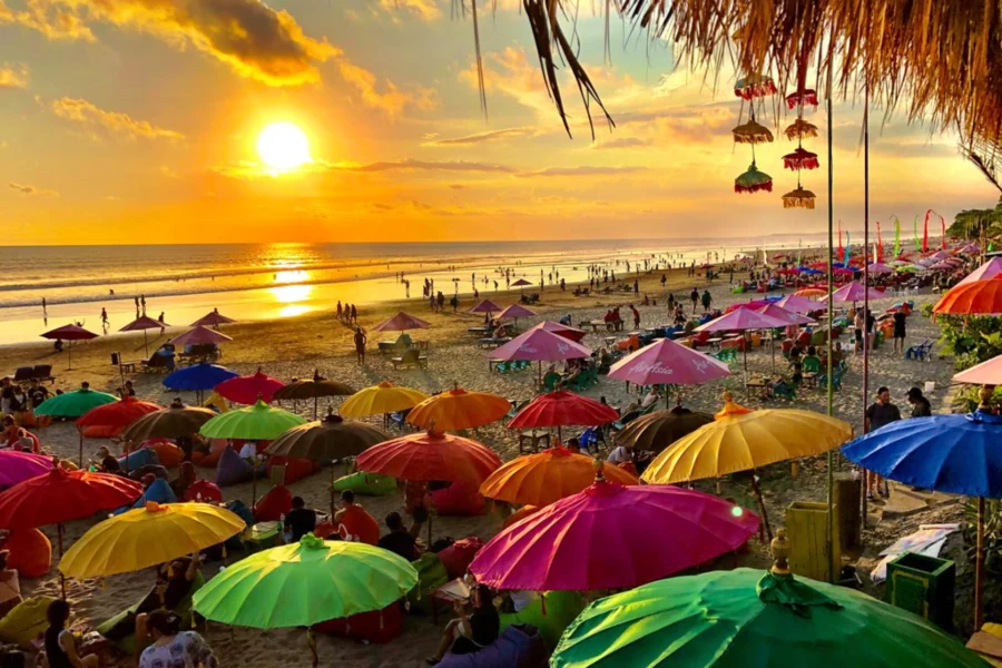 Crowded tropical beach at sunset with colorful umbrellas, people relaxing, and golden ocean waves.