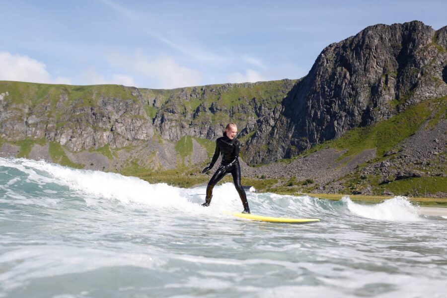 Surfer in black wetsuit on yellow surfboard riding a small coastal wave near cliffs.
