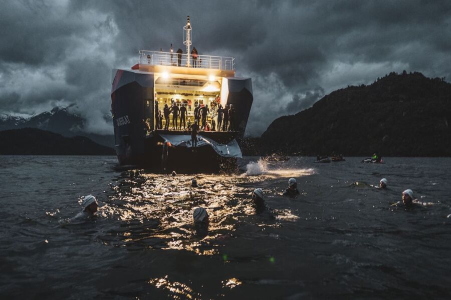 Swimmers navigate dark waters near an illuminated ship and rugged mountains, under stormy skies.
