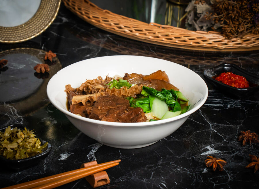 Side view of a bowl of braised beef noodles with leafy greens and chopsticks, isolated on a dark background, representing Taiwanese food